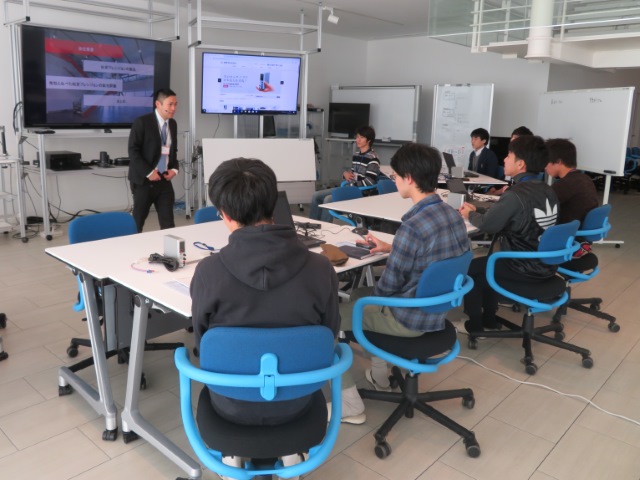 参加者が椅子に座って大型モニタの前で説明している社員の話を聞いている写真です。